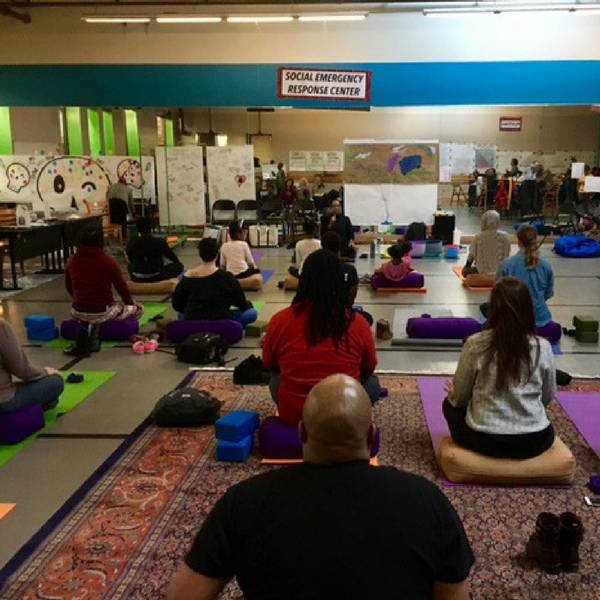 Thisis an image looking at the back of several people doing yoga. There is a large mirror in the front. On the mirror is a sign that says, "Social Emergency Response Center".